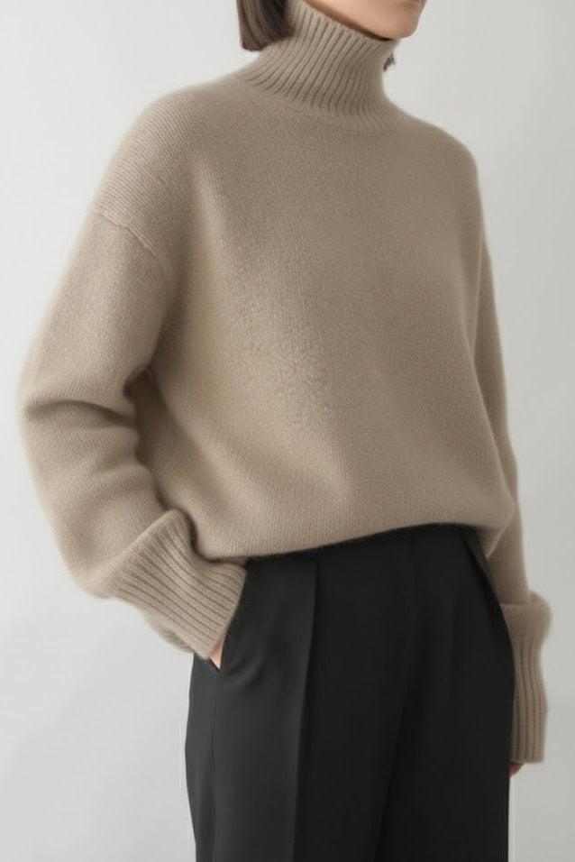 Person wearing a beige turtleneck sweater and dark pants on a plain background