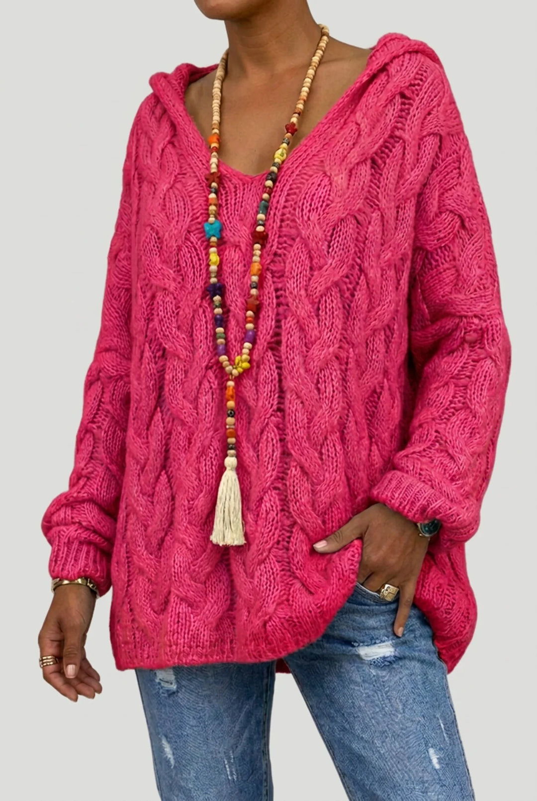 A woman wearing a bright pink cable-knit hooded sweater, blue jeans, and a long beaded tassel necklace, shown from the neck down against a light gray background.